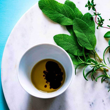 Olive oil with balsamic vinegar in a white bowl beside fresh sage, rosemary, and thyme herbs