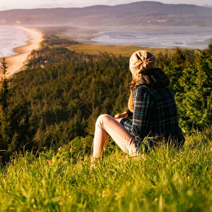 Woman sitting on a hillside overlooking a scenic coastal landscape with green forests and sandy beach