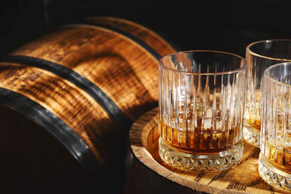 Two crystal glasses of whiskey with ice resting on a wooden barrel in warm amber light