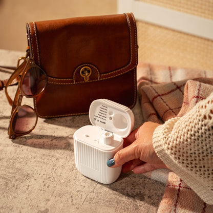 A hand with blue nail polish holds an open white portable scent diffuser with a ribbed case. In the background, a brown leather pouch, sunglasses, and a plaid blanket are arranged on a textured surface.