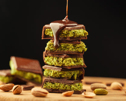 Stacked pistachio chocolate bars with melted chocolate drizzle and pistachios scattered on wooden surface
