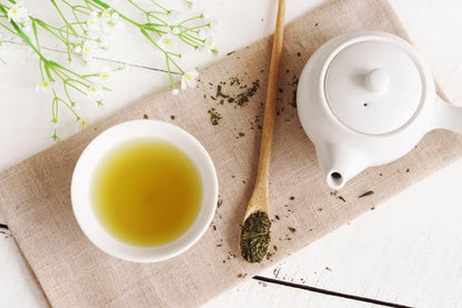 Green tea brewed in a white bowl beside a teapot and wooden spoon with loose green tea leaves