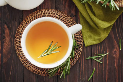 Rosemary herbal tea in white cup on woven mat, fresh rosemary sprigs on dark wood table