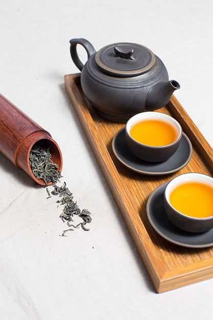 Green tea leaves spilling from bamboo container beside dark ceramic teapot and cups with golden tea