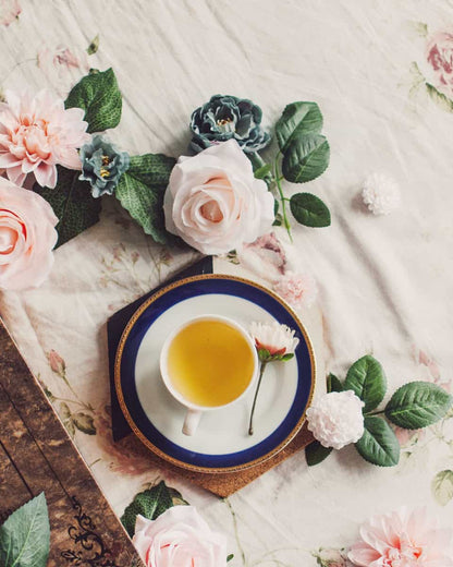 White tea cup with green tea on floral backdrop surrounded by pink roses and blue flowers